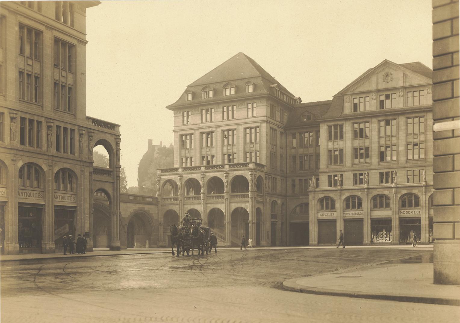 Ein historisches Schwarz-Weiss-Foto, das einen Platz in einer Stadt zeigt. Mehrere grosse, mehrstöckige Gebäude mit Arkaden und Schaufenstern umgeben den Platz. In der Mitte ist eine Pferdekutsche zu sehen. 