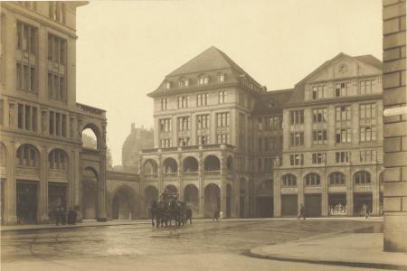 Ein historisches Schwarz-Weiss-Foto, das einen Platz in einer Stadt zeigt. Mehrere grosse, mehrstöckige Gebäude mit Arkaden und Schaufenstern umgeben den Platz. In der Mitte ist eine Pferdekutsche zu sehen. 