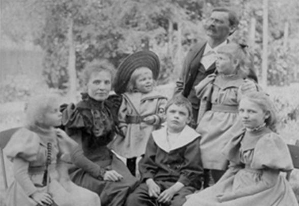 Ein historisches, schwarz-weisses Porträt einer Familie. Die Familie sitzt draussen, umgeben von Bäumen und Gras. 