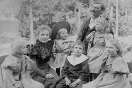 Ein historisches, schwarz-weisses Porträt einer Familie. Die Familie sitzt draussen, umgeben von Bäumen und Gras. 