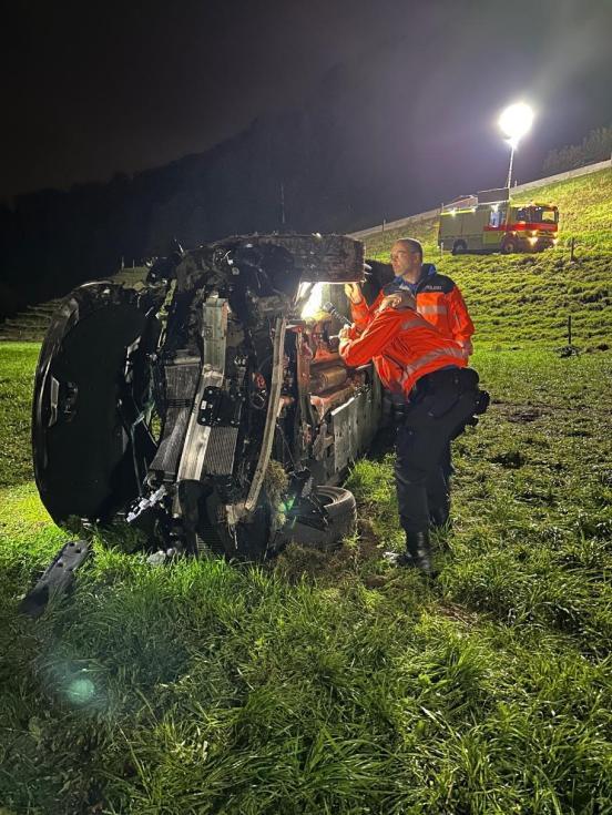 Unfallfahrzeug liegt auf der Seite in einer Wiese und zwei Polizisten inspizieren die Fahrzeugunterseite. Im Hintergrund steht ein Feuerwehrauto mit einer grossen Balonleuchte, die die Nacht ausleuchtet.