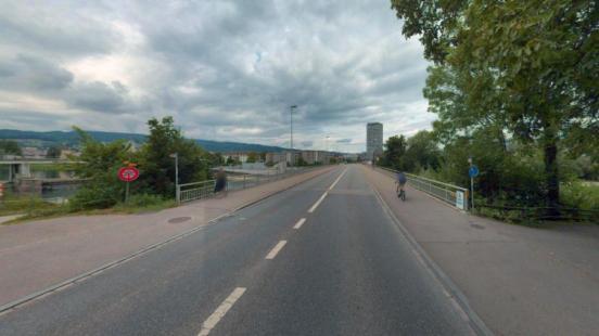 Asphaltierte Brücke mit Fahrbahn, Fussgänger- und Radweg unter bewölktem Himmel, einem hohen Gebäude in der Ferne, und Bäumen an den Seiten.