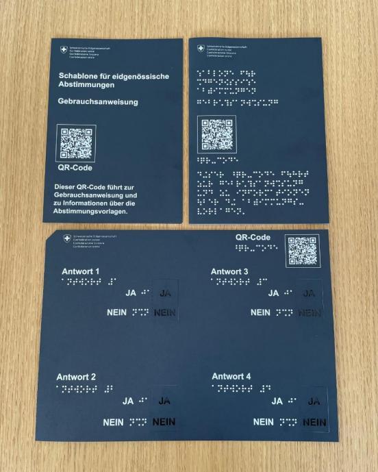 Auf einem braunen Holztisch sind die Abstimmungsschablone und je eine Gebrauchsanweisung in Druckschrift und eine Brailleschrift aufgelegt. Die Abstimmungsschablone und die Gebrauchsanweisungen sind aus schwarzem Karton gefertigt. Bedruckt sind sie mit weisser grosser Druckschrift und mit Brailleschrift. Zudem ist sowohl auf der Schablone als auch auf den Gebrauchsanweisungen ein QR-Code aufgedruckt.