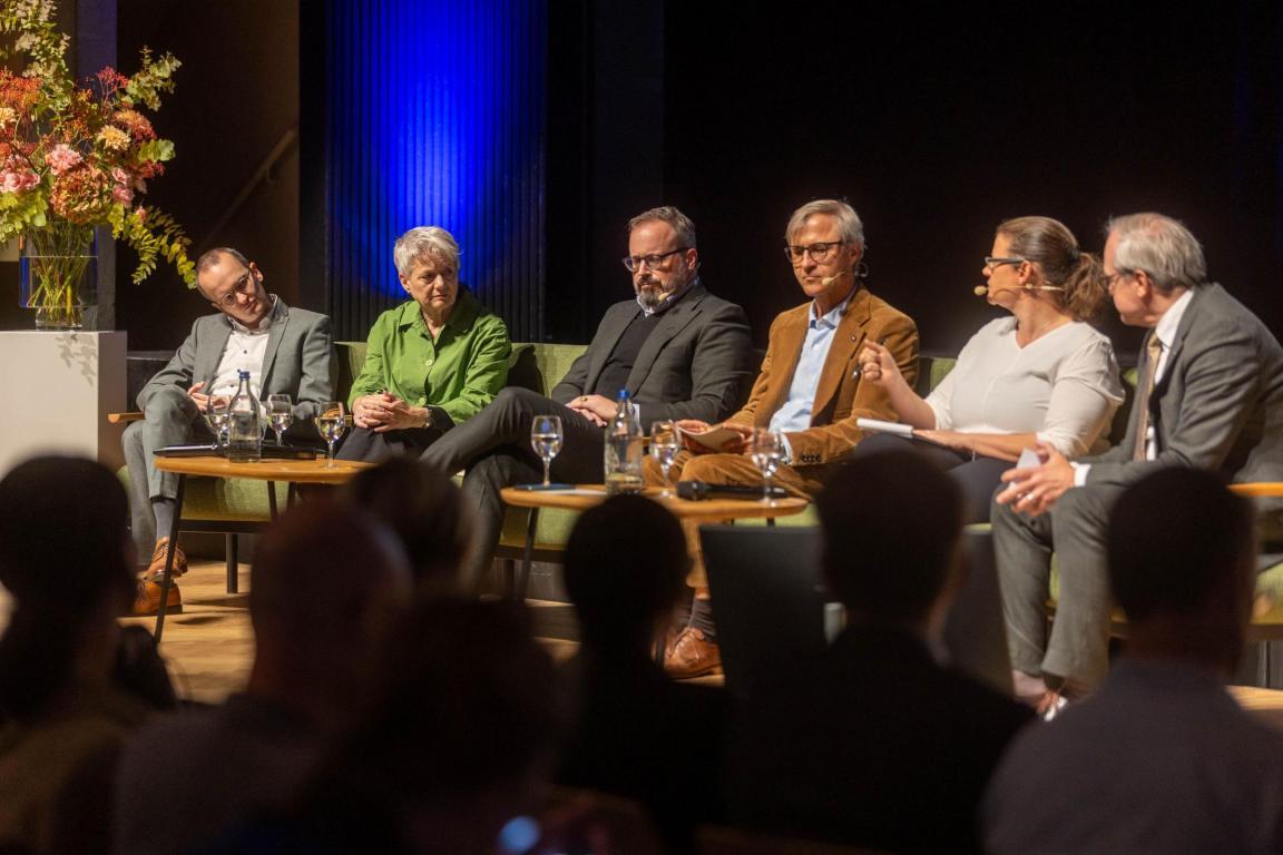 Die sechs Podiumsgäste sitzen in einer Reihe auf der Bühne.