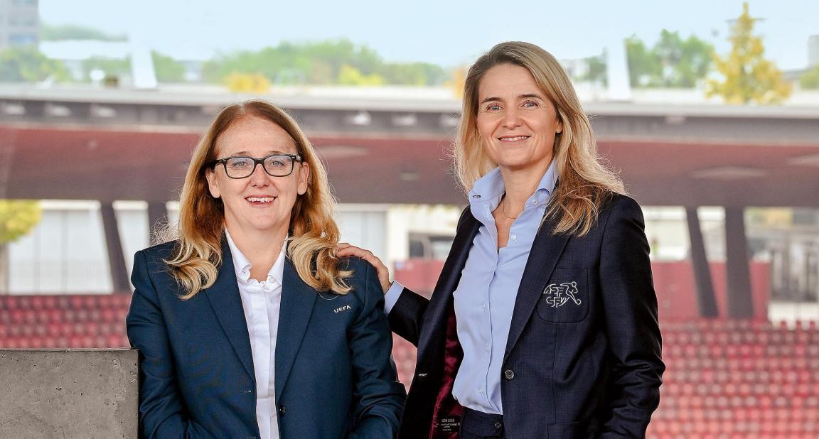 Zwei lächelnde Frauen mit blonden, langen Haaren und in je dunkelblauem Anzug in einem leeren Fussballstadion mit roten Sitzen.