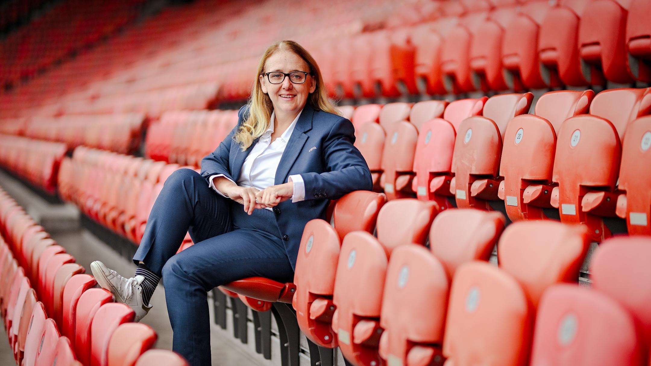 Blonde, lächelnde Frau mit Brille im Anzug auf rotem Stadionsitz mitten in me