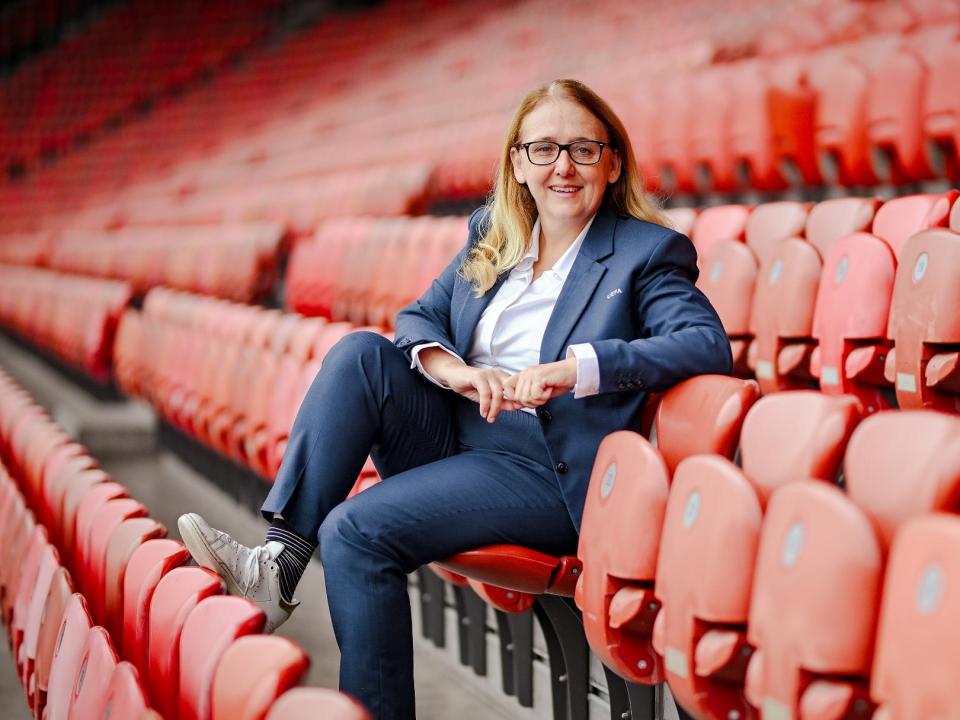 Blonde, lächelnde Frau mit Brille im Anzug auf rotem Stadionsitz mitten in me