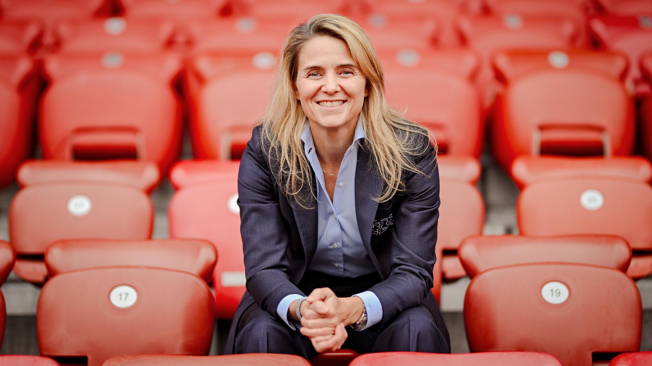 Lächelnde Frau mit blonden Haaren und dunklem Anzug sitzt auf einer leeren, roten Stadiontribüne.