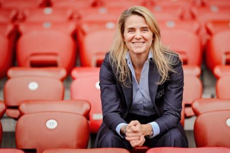 Lächelnde Frau mit blonden Haaren und dunklem Anzug sitzt auf einer leeren, roten Stadiontribüne.