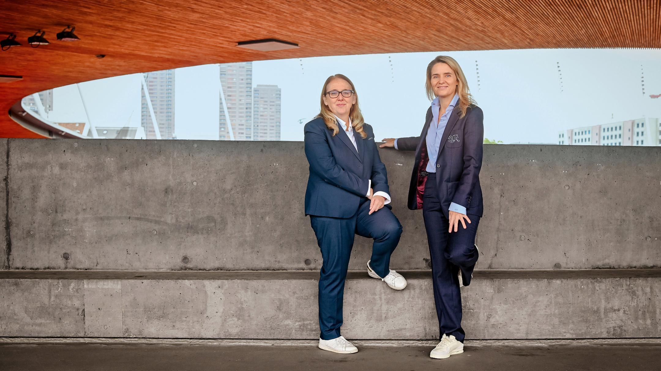Zwei Frauen in dunklen Anzügen und weissen Sneakern posieren in einem Fussballstadion vor einer grauen Betonmauer mit  Holzdecke.