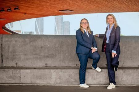 Zwei Frauen in dunklen Anzügen und weissen Sneakern posieren in einem Fussballstadion vor einer grauen Betonmauer mit  Holzdecke.
