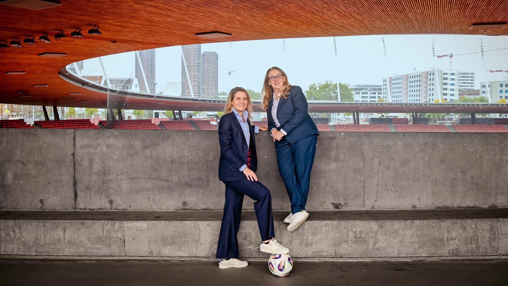 Zwei Frauen in dunklen Anzügen und weissen Sneakern posieren mit Ball in einem Fussballstadion vor einer grauen Betonmauer mit  Holzdecke.