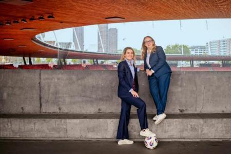 Zwei Frauen in dunklen Anzügen und weissen Sneakern posieren mit Ball in einem Fussballstadion vor einer grauen Betonmauer mit  Holzdecke.