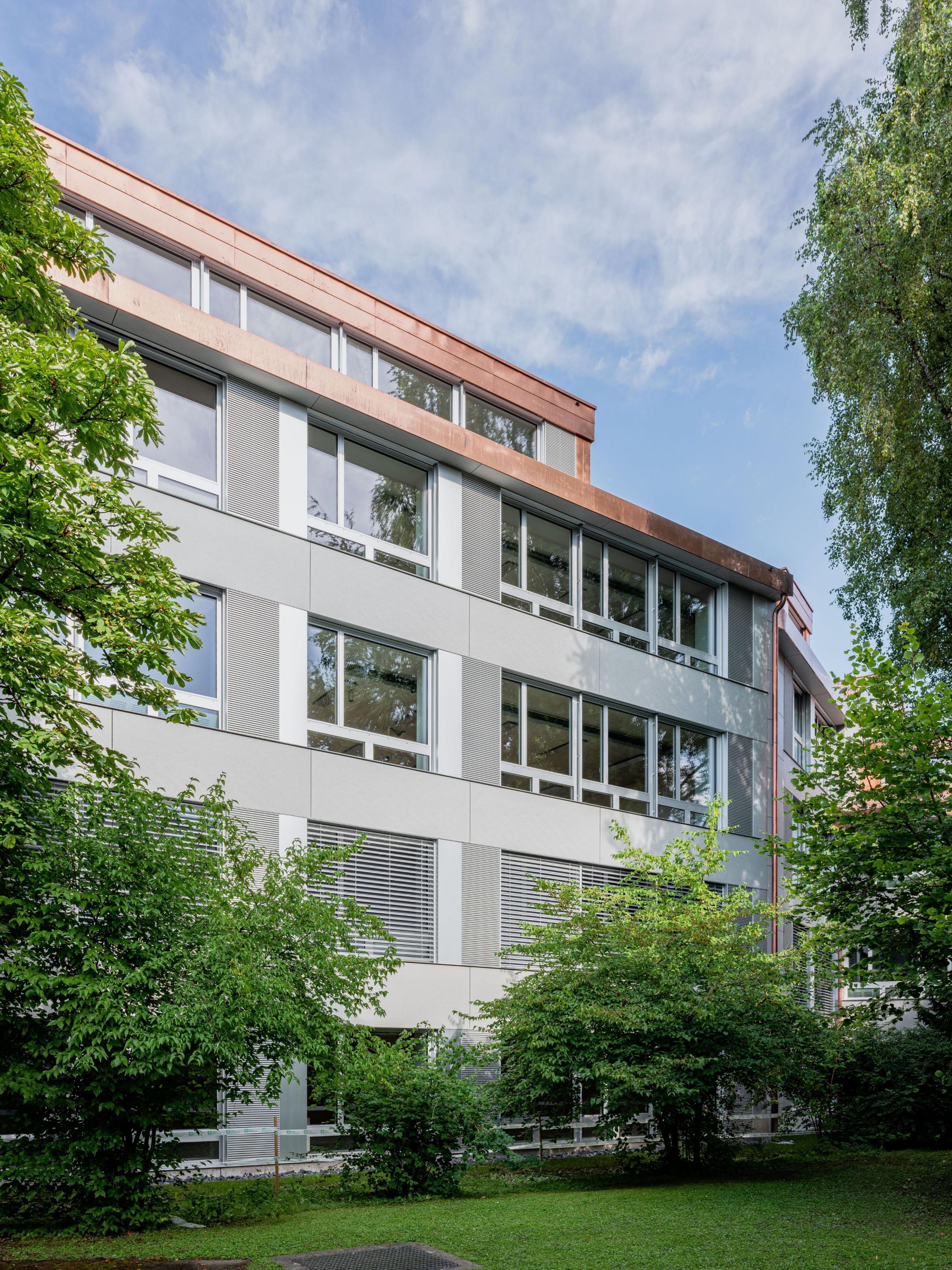 Aussenansicht des Gebäudes mit einheitlich gestalteter Fassade in Grautönen und Bäumen rundherum.
