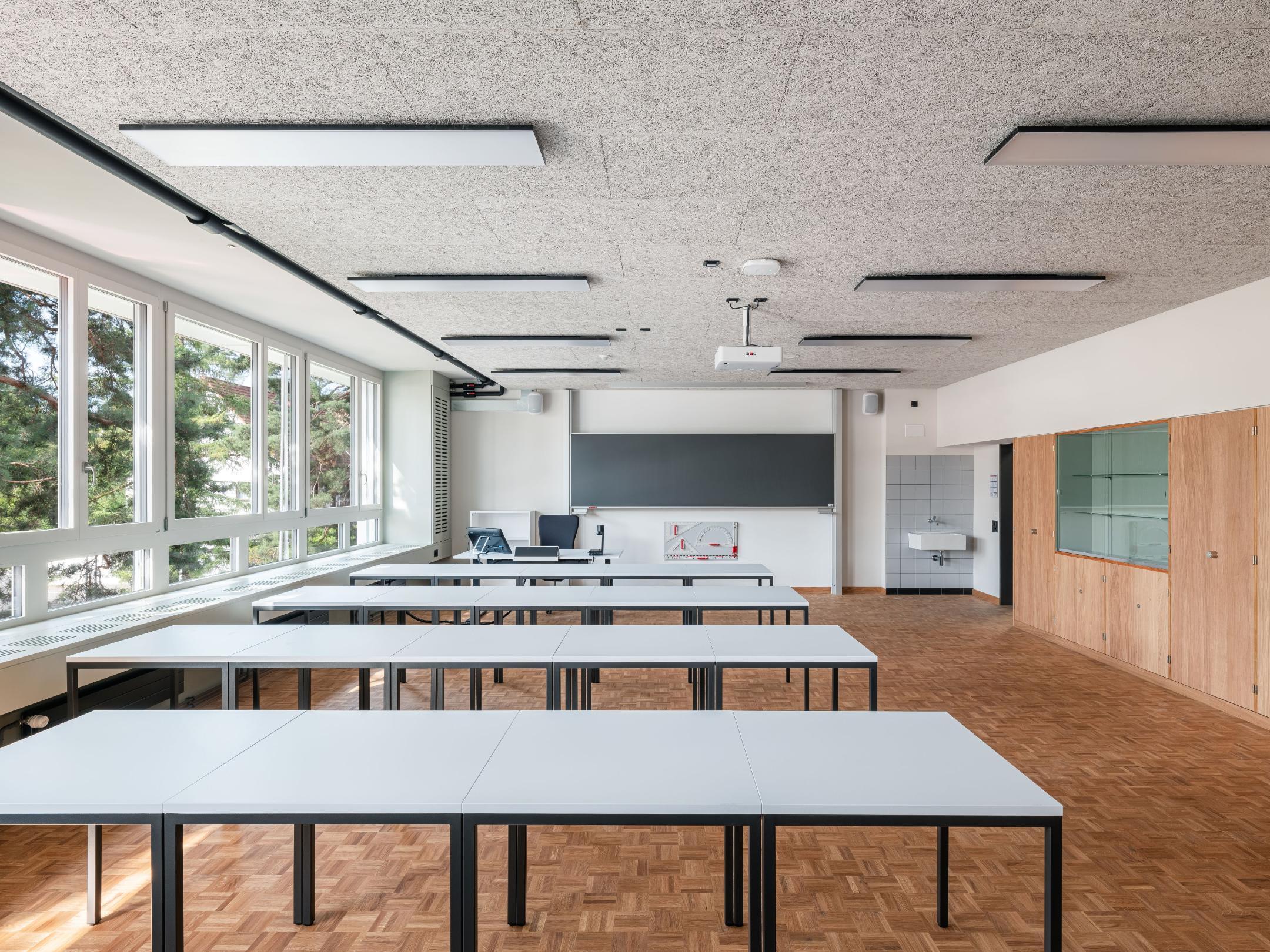 Klassenzimmer mit vier Tischreihen, Fensterfront auf der linken Seite, Decke aus Holzwollplatten und Parkettboden. 
