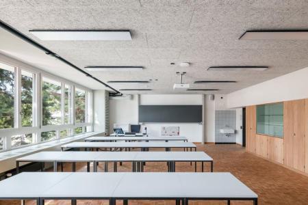 Klassenzimmer mit vier Tischreihen, Fensterfront auf der linken Seite, Decke aus Holzwollplatten und Parkettboden. 