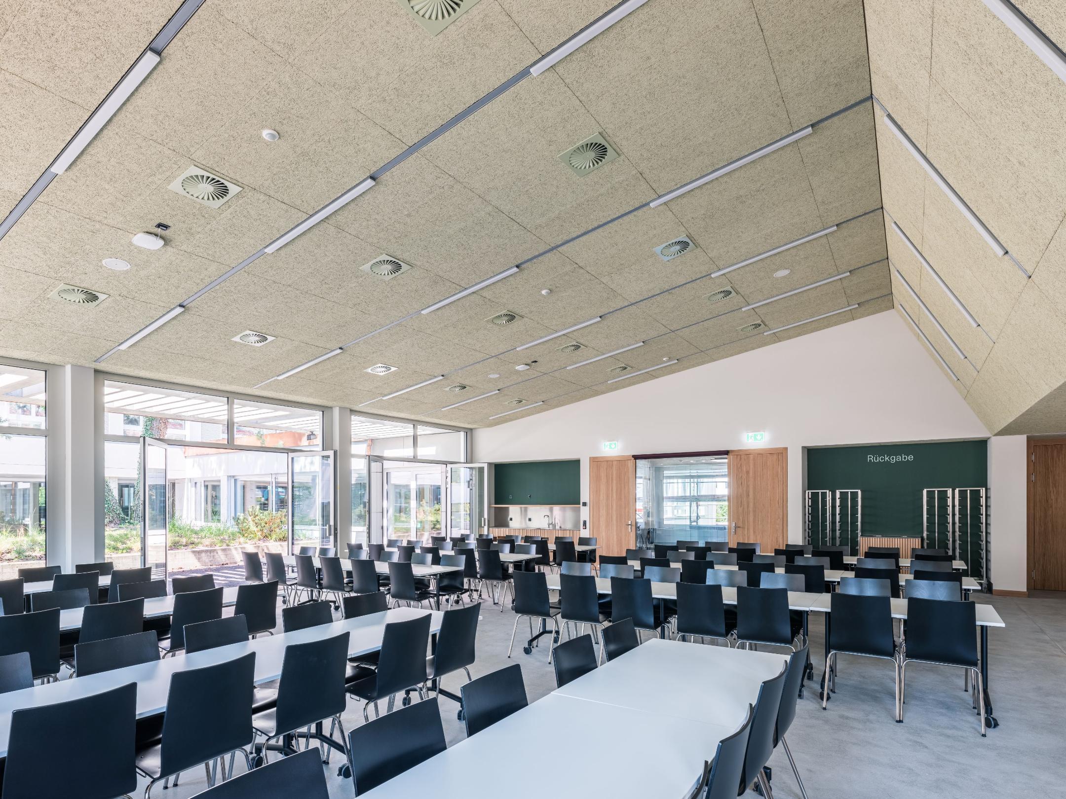 Blick in die Mensa mit hohen Decken, grosser Fensterfront und diversen Tischen mit Stühlen.