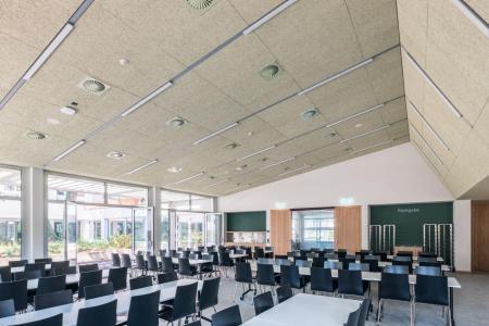 Blick in die Mensa mit hohen Decken, grosser Fensterfront und diversen Tischen mit Stühlen.