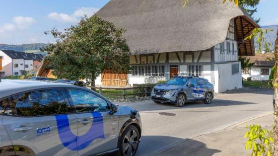 Zwei Personenfahrzeuge von Nissan, die auf dem Autodach mit Sensoren zum automatisierten Fahren ausgerüstet sind, kreuzen sich auf der Landstrasse in Hüttikon. Im Hintergrund ist ein Strohdachhaus zu sehen.