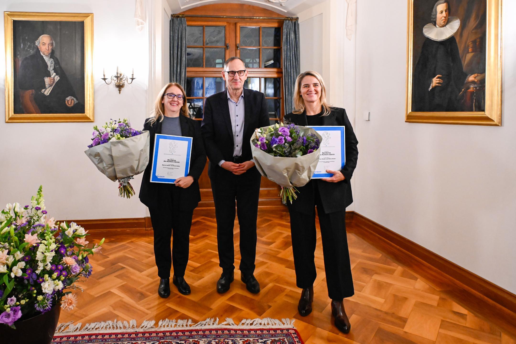 Zwei Frauen und ein Mann in Anzug und Brille in einem festlichen Saal halten je eine Urkunde und einen Blumenstrauss in der Hand.