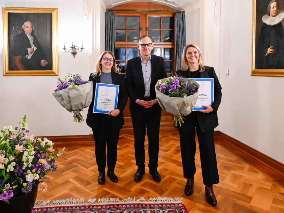 Zwei Frauen und ein Mann in Anzug und Brille in einem festlichen Saal halten je eine Urkunde und einen Blumenstrauss in der Hand.