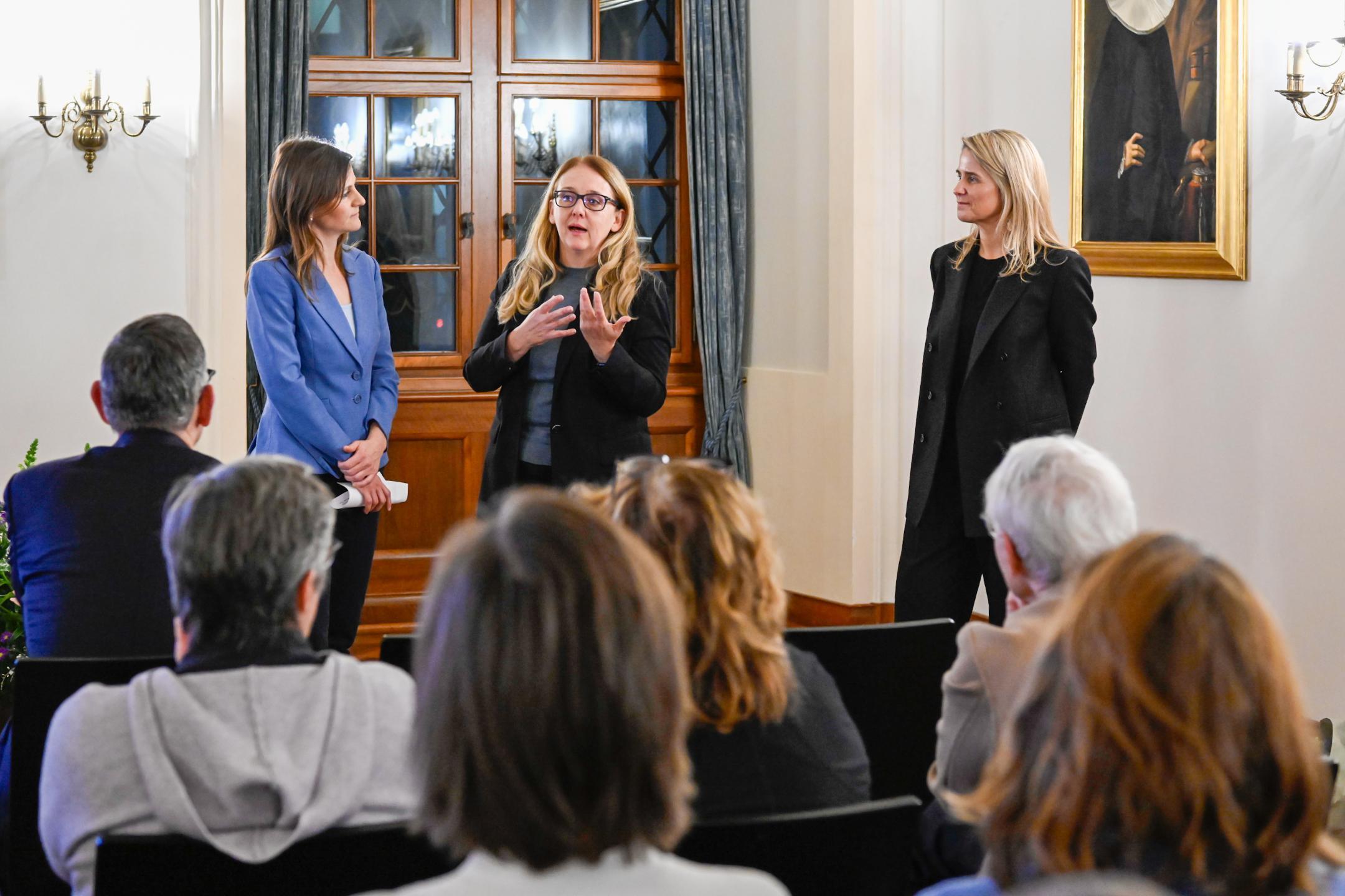 Eine Frau mit langen, braunen Haaren spricht mit zwei blonden Frauen, die links und rechts von ihr stehen.