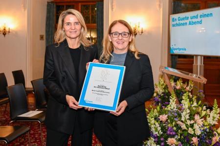 Zwei Frauen mit blonden Haaren posieren mit Urkunde und Blumen in der Hand.
