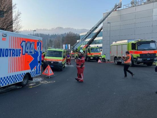 Feuerwehr- und Rettungsdienstfahrzeuge auf einer Strasse. Im Hintergrund zwei Drehleitern der Feuerwehr an einem Gebäude.