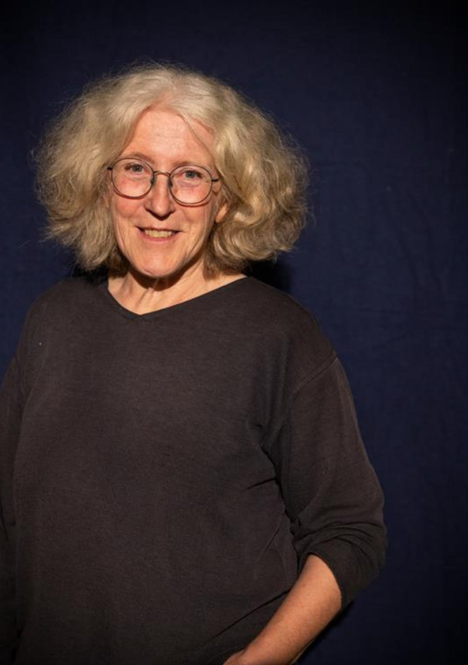 Ursula Bienz, eine Frau mit halblangem grauem Haar. Sie trägt einen schwarzen Pullover und steht vor einem dunklen Hintergrund.