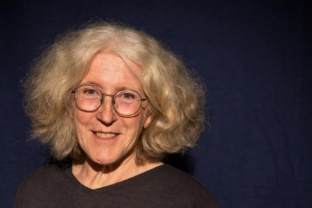 Ursula Bienz, eine Frau mit halblangem grauem Haar. Sie trägt einen schwarzen Pullover und steht vor einem dunklen Hintergrund.