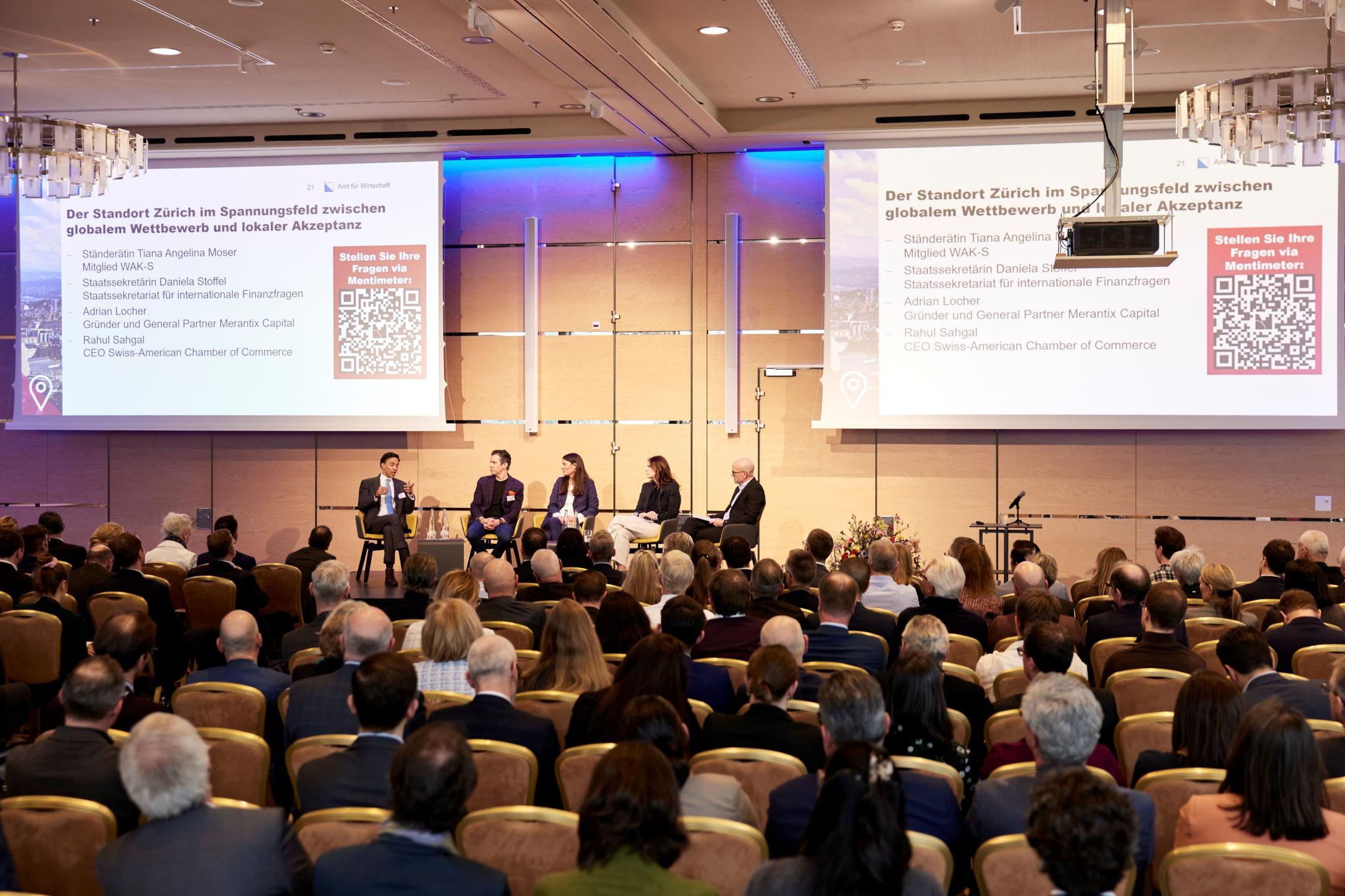 Fünf Personen sitzen auf einer Bühne und sprechen miteinander, im Saal sitzen Zuschauer und hören zu