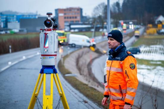 Polizist mit einem Gerät auf einer Unfallstelle. Im Hintergrund in der Unschärfe befinden sich zwei Unfallfahrzeuge