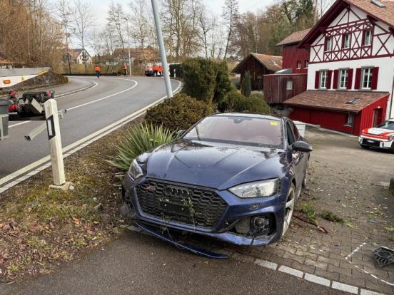 Das verunfallte Auto steht neben der Strasse, auf einem Privatsträsschen, parallel zur öffentlichen Strasse in Kollisionsendstellung. Im Hintergrund ein Polizeiauto und ein Riegelhaus.