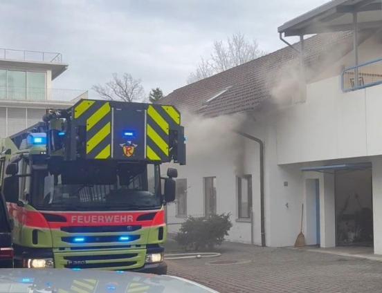 Ein Feuerwehrauto steht mit eingeschaltetem Blauchlicht voreinem Haus aus welchem Rauch dringt.