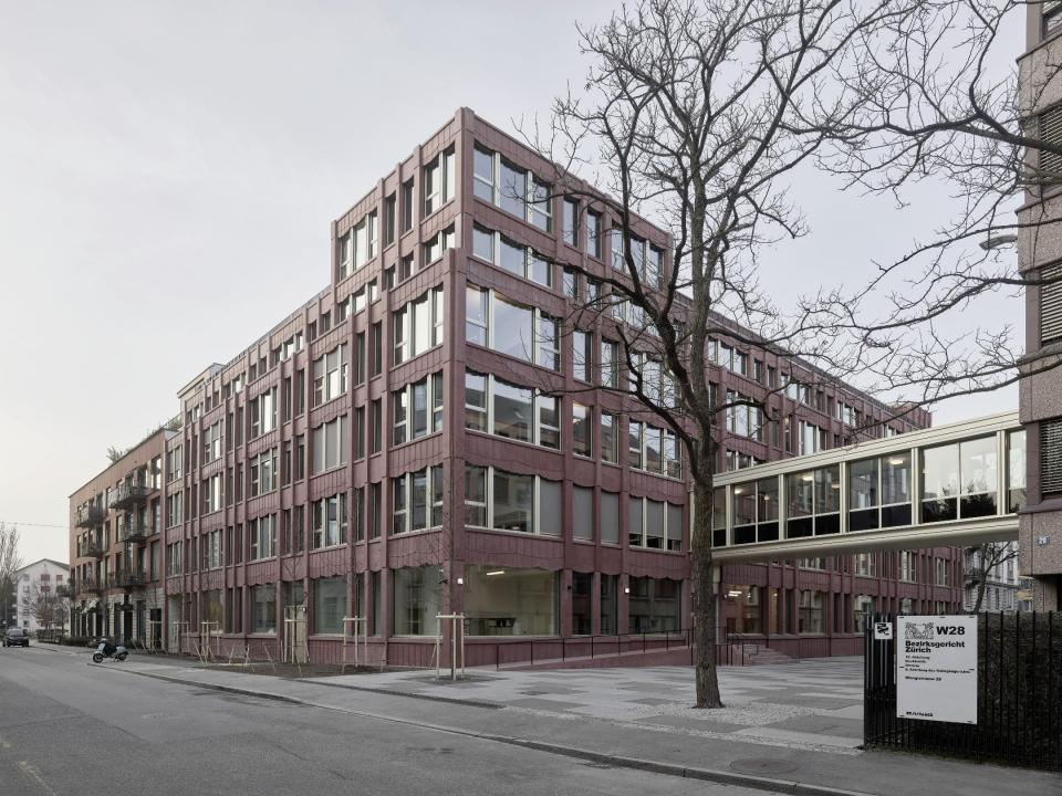 Gerichtsgebäude mit roter Fassade und horizontalen Fenstern, dazwischen ein verglaster Übergang zum Nachbargebäude. Am Zaun ein Schild mit der Aufschrift «Bezirksgericht Zürich W28».