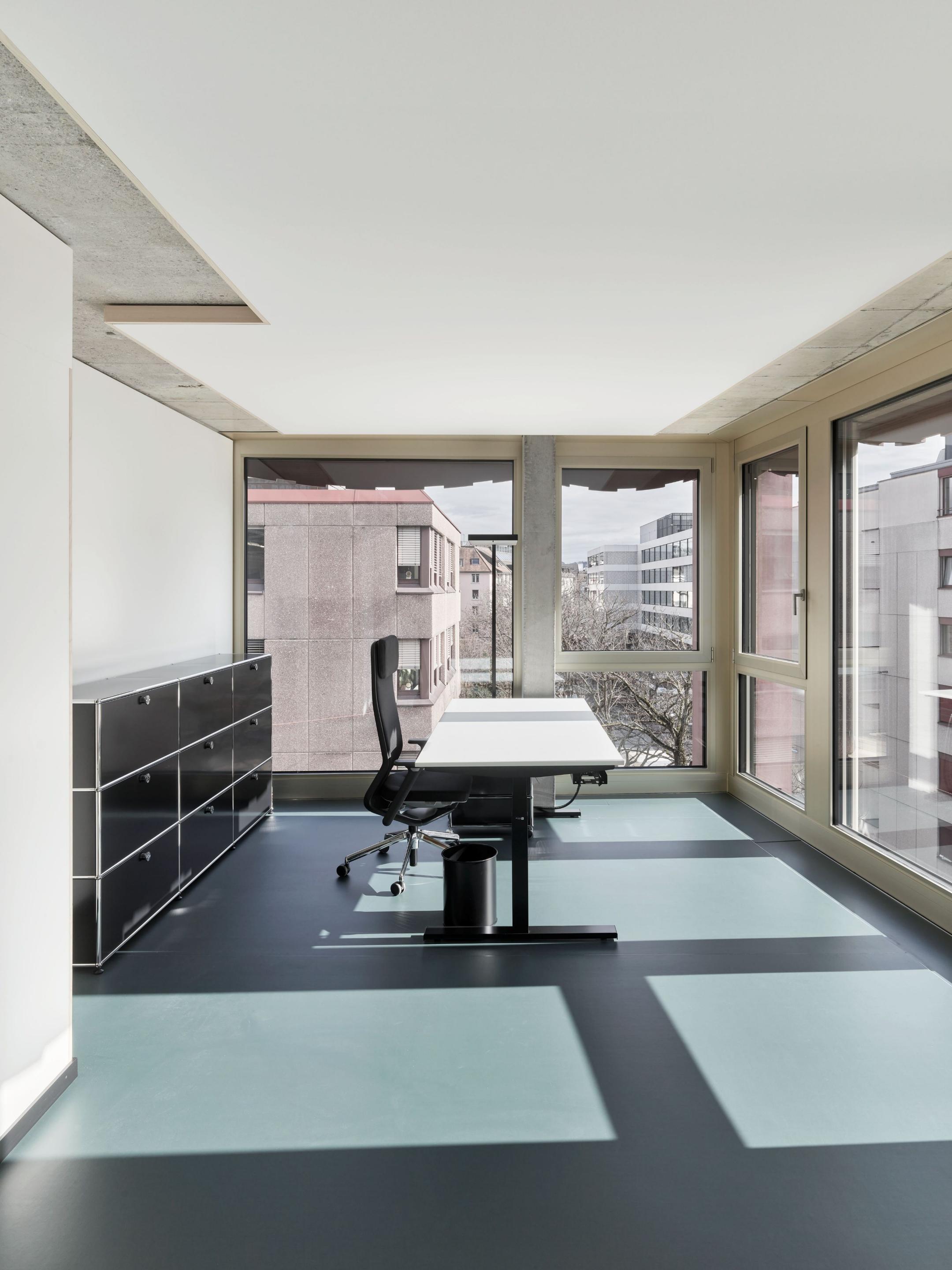 Modernes Bürozimmer mit hellblau-grauem Boden, einem schwarzen Büromöbel, weissem Schreibtisch und Bürostuhl vor grossem Fenster. 