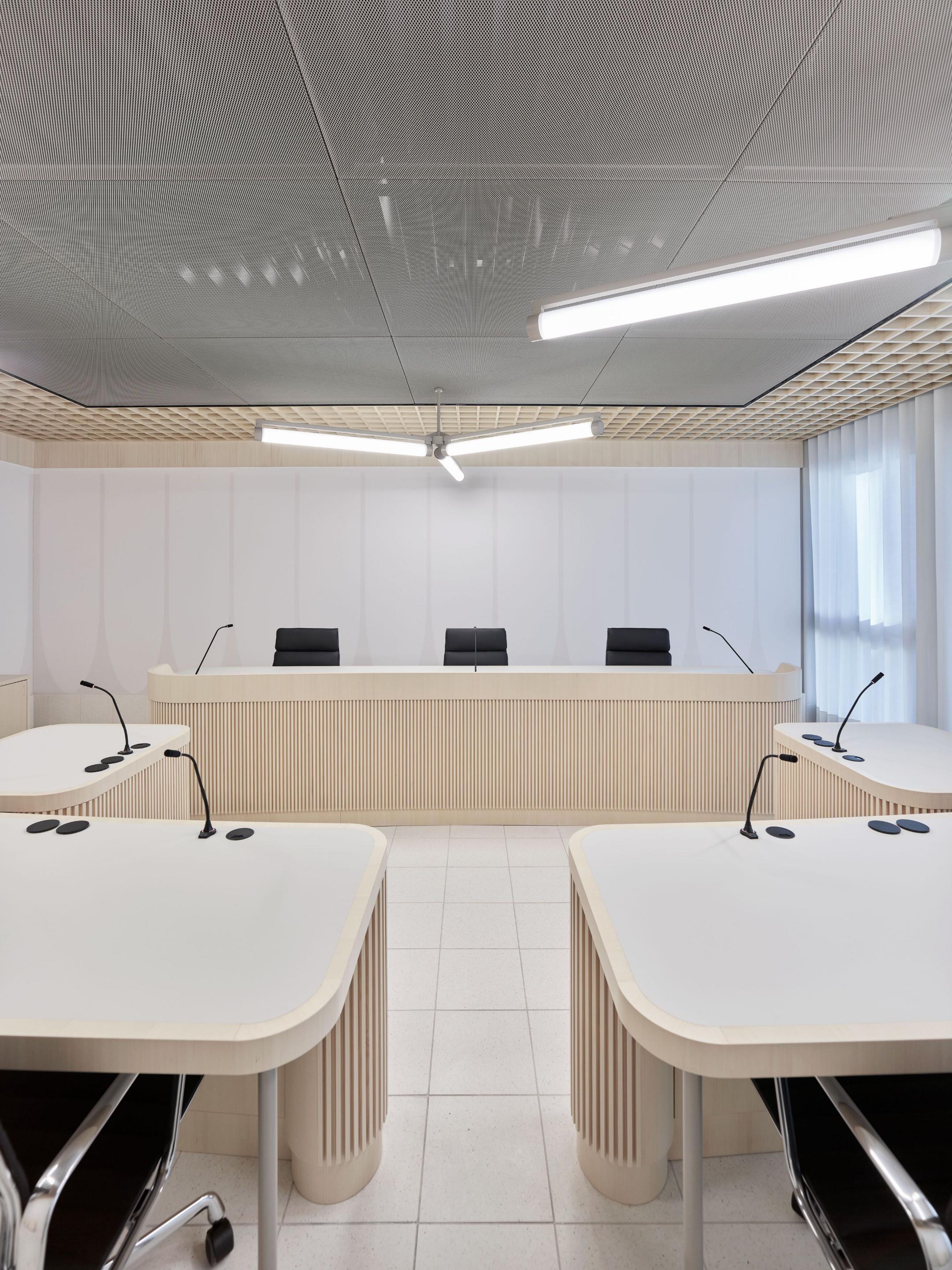 Moderner Gerichtssaal mit zwei hellen, geschwungenen Tischreihen, drei schwarzen Bürostühlen hinter einem langen Holzpult und weiss-silberner Decke.
