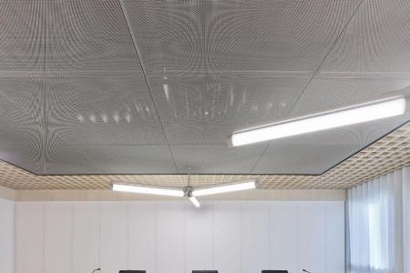 Moderner Gerichtssaal mit zwei hellen, geschwungenen Tischreihen, drei schwarzen Bürostühlen hinter einem langen Holzpult und weiss-silberner Decke.