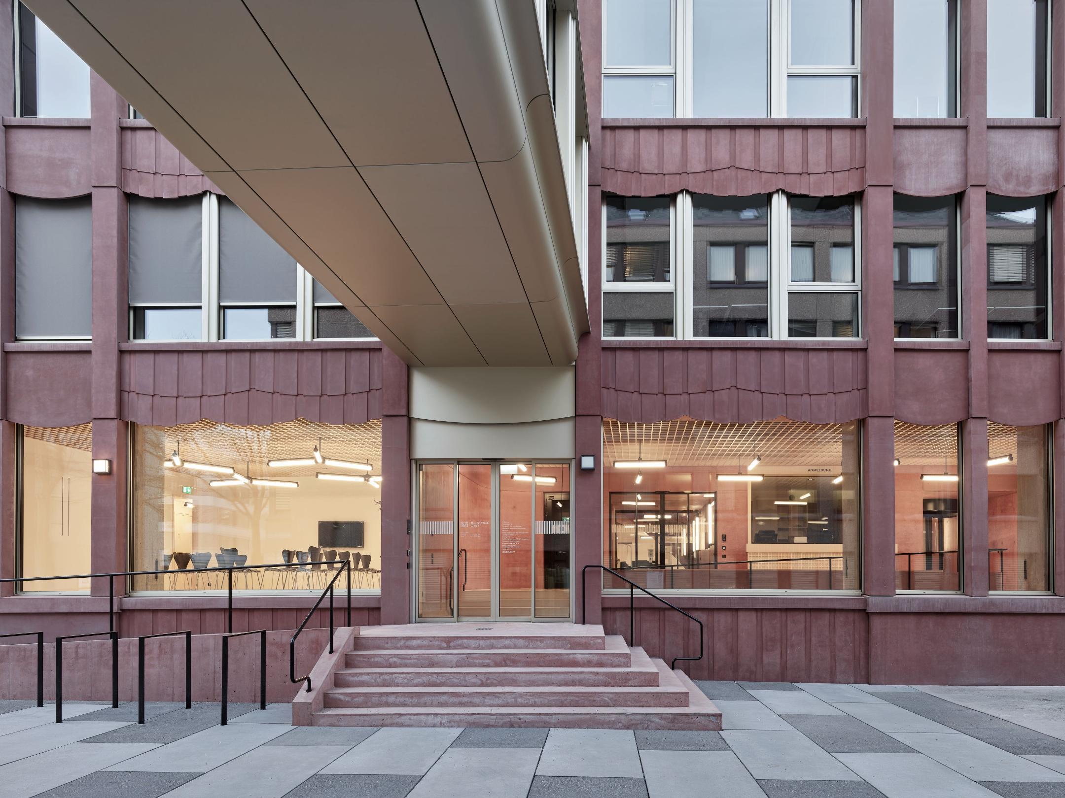 Eingangsbereich eines modernen Gebäudes mit roter Fassaden, einer Rampe, Treppen und einem überdachten Übergang.