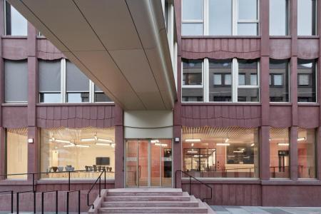 Eingangsbereich eines modernen Gebäudes mit roter Fassaden, einer Rampe, Treppen und einem überdachten Übergang.