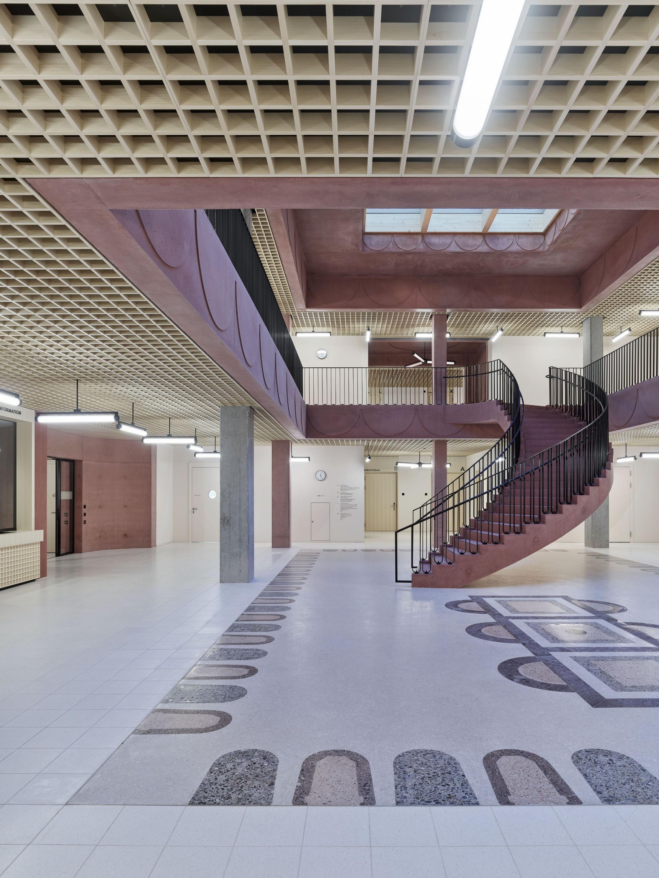 Grosser Empfangs- oder Wartebereich mit rötlichen Wänden und Betonsäulen, einer geschwungenen Treppe und einem holzgerasterten Deckenraster.