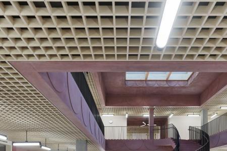 Grosser Empfangs- oder Wartebereich mit rötlichen Wänden und Betonsäulen, einer geschwungenen Treppe und einem holzgerasterten Deckenraster.