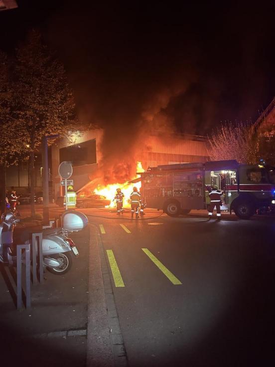 Im Hintergrund zwei brennende Auots, im Vordergrund ein grosses Feuerwehrautos und mehrere Feuerwehrmänner.