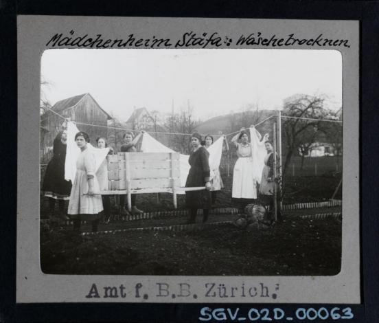 Man sieht junge Frauen, die Wäsche an eine Leine aufhängen. Das Bild ist eine Schwarz-Weiss-Fotografie. 