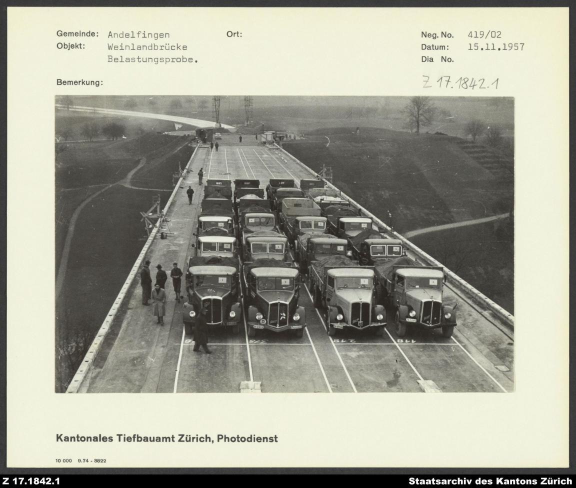 Auf dem Schwarzweissbild sieht man alte Lastwagen aufgereiht auf einer Autobahnbrücke, daneben stehen einige Männer