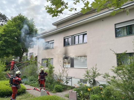Feuerwehrleute vor einem Mehrfamilienhaus, aus dessen Fenster Rauch emporsteigt