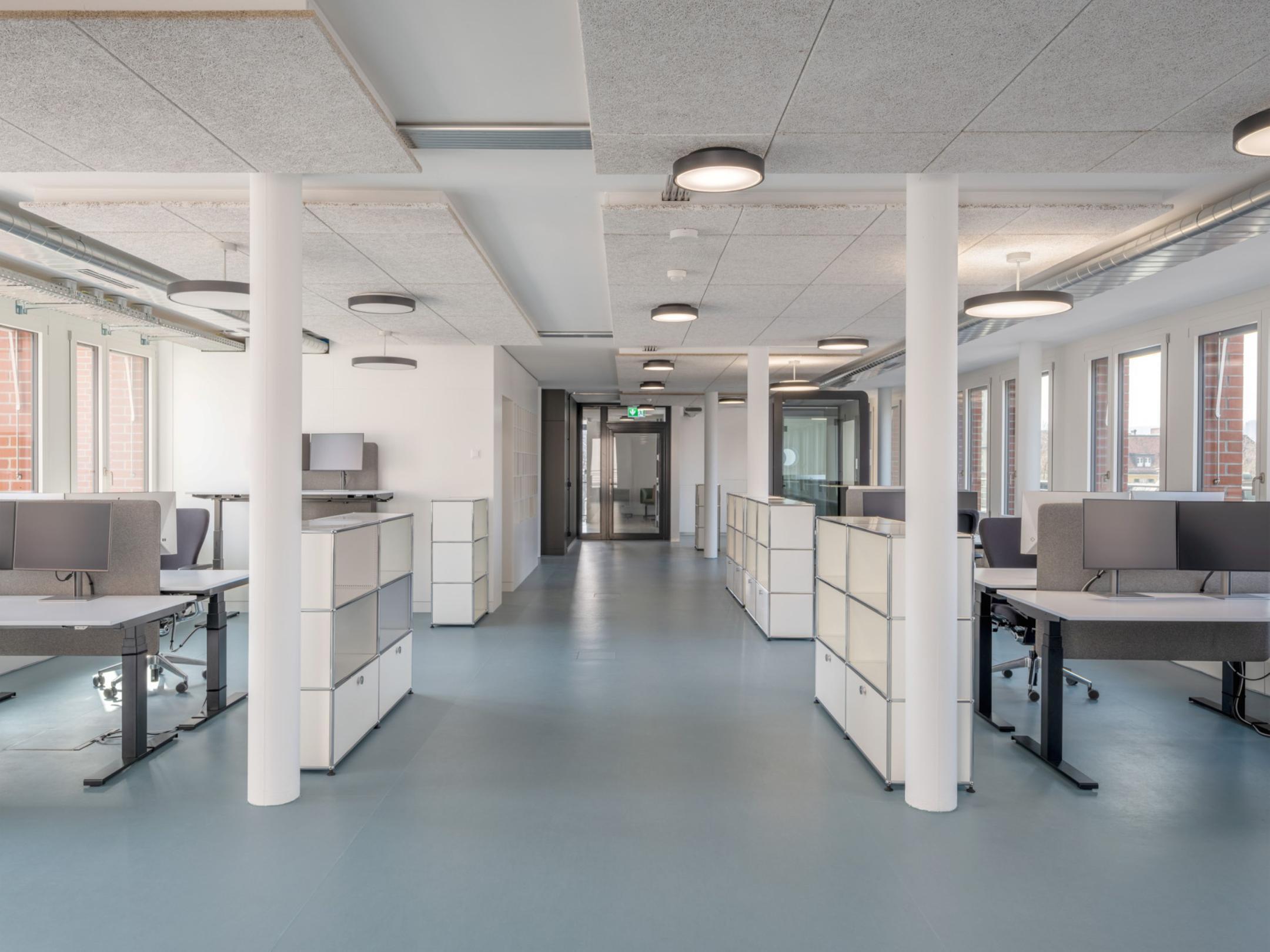 Moderner Grossraumbüro mit Computertischen und Schränken, helle Lichtdecken und Fenster mit Ziegelmauerwerk draussen.