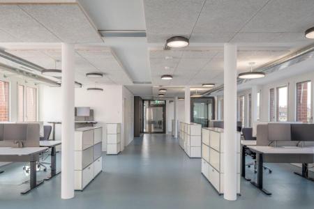 Moderner Grossraumbüro mit Computertischen und Schränken, helle Lichtdecken und Fenster mit Ziegelmauerwerk draussen.