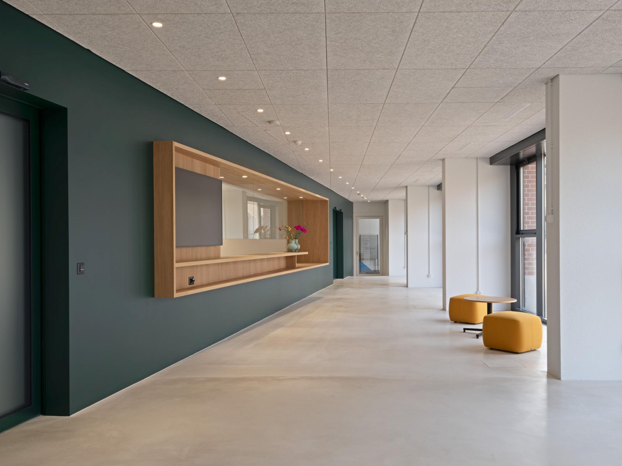 Langer Flur mit dunkelgrüner Wand, Holzalmosen, gelben Sitzhocker und einem Beistelltisch.