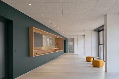 Langer Flur mit dunkelgrüner Wand, Holzalmosen, gelben Sitzhocker und einem Beistelltisch.